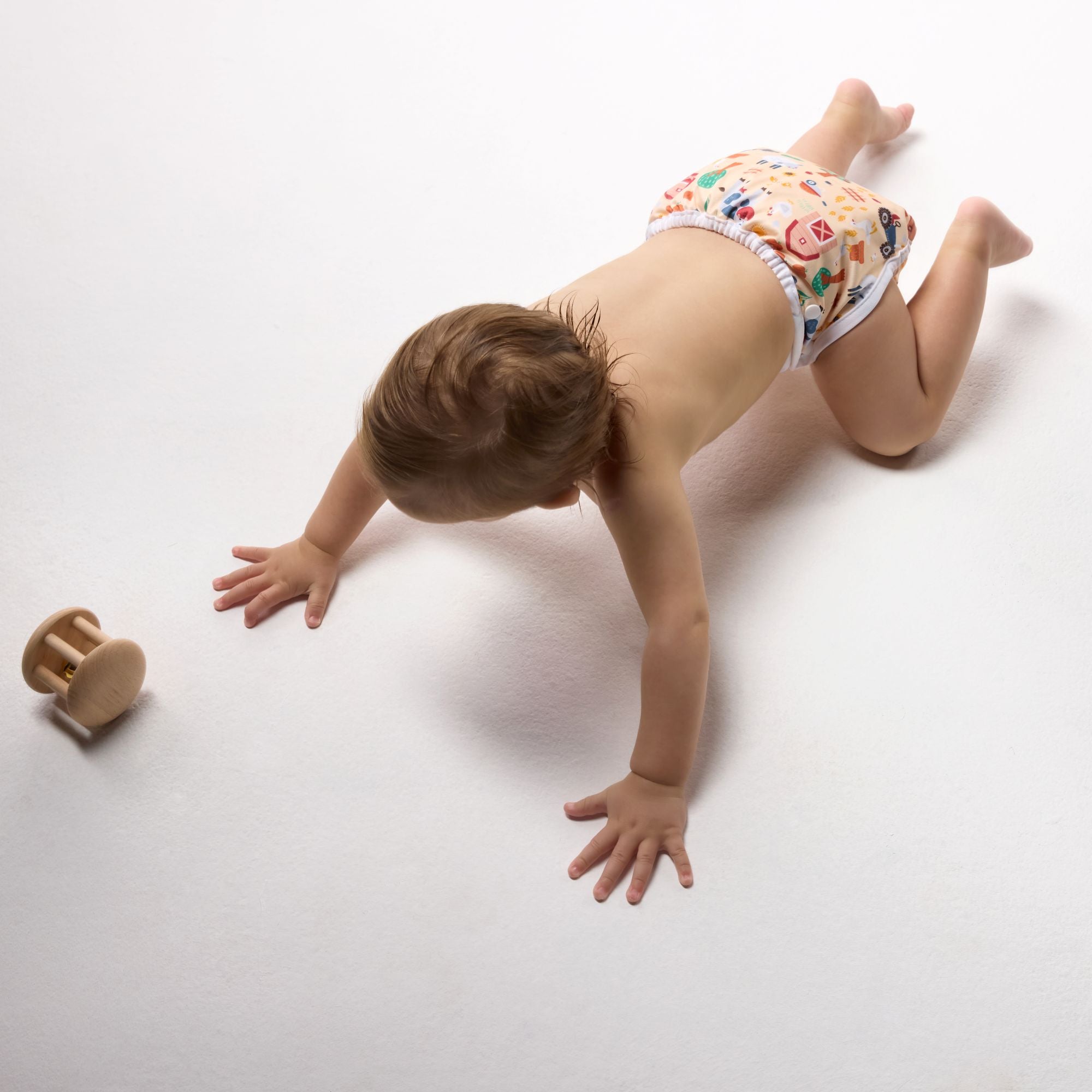 Baby in a colorful Bonjour Babies diaper crawling on a white surface with a wooden toy nearby.